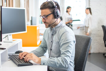 Handsome male customer support phone operator with headset working on desktop computer in office.