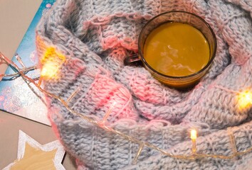 A cup of coffee with milk on a table with knitten blue plaid and garland. Atmosphere of winter holidays.