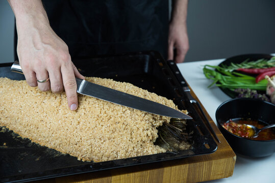 Chief cook is breaking salt and carving fish baked in salt crust. Seafood cooking. Salmon or seabass under salt crust with vegetables.