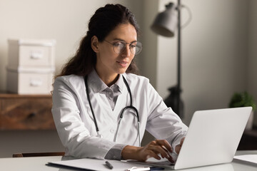 Focused female GP or nurse in white medical uniform look at laptop screen consult client or patient online. Successful woman doctor work on computer in modern clinic or hospital. Medicine concept.