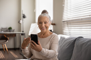 Smiling modern Caucasian senior woman look at cellphone screen have video call on gadget at home....