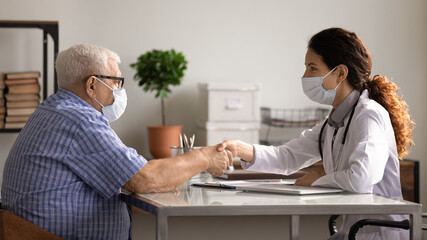 Female doctor and old male patient in protective facemasks handshake close health insurance deal in hospital. Woman GP in facial masks from covid-19 shake hand greeting with mature client in clinic.