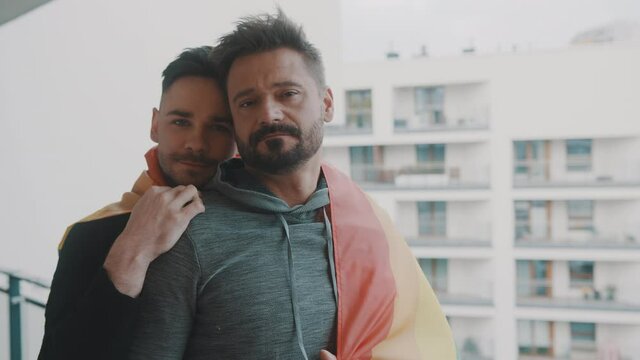 Young excited gay male couple hugging on the balcony covered with rainbow flag. . High quality 4k footage