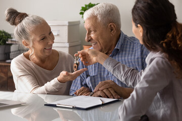 Female real estate agent give keys to new home to excited elderly couple clients. Woman realtor or broker congratulate happy overjoyed mature spouses with house purchase. Ownership concept.