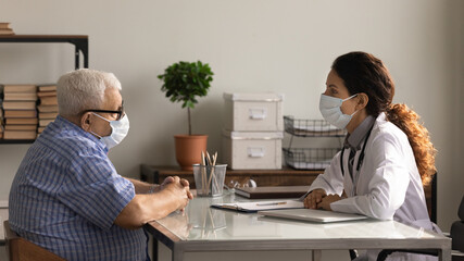 Female doctor in facial mask have consultation with old senior patient in hospital. Woman GP or therapist consult mature male client during corona virus pandemics quarantine. Covid-19 concept.