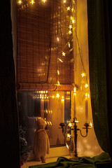 
hygge, decoration and christmas concept - candles burn in lanterns on the windowsill and festive garland at home