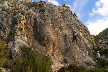 Fototapeta premium Young girl climber in the middle of a rock wall while her partner belays her from below