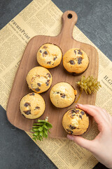 Above view of hand holding one of small cupcakes with accessories on wooden cutting board on newspaper on dark background