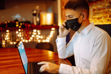 Young man wearing medical face mask working on laptop computer  and speaks on the phone at home during winter holiday. The concept of preventing the spread of the epidemic coronavirus. Remote work. 