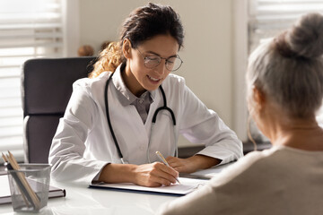 Smiling young woman GP write prescription to mature patient at consultation in hospital. Female...