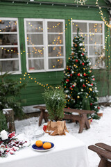 Plates and glasses are placed on the dining table, the table is on the street. There is snow and a Christmas tree is decorated. The country house is decorated with a garland. Christmas dinner outdoors