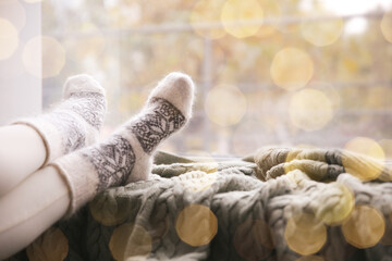 Woman wearing warm socks on knitted plaid near window, closeup. Space for text