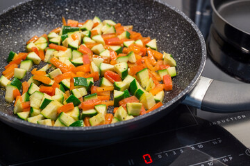 Diced carrots, paprika, and zucchini are sautéed in a black, non-stick frying pan on a modern induction stove