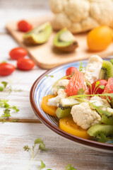 Vegetarian salad of cauliflower cabbage, kiwi, tomatoes, microgreen sprouts on gray wooden background. Side view, selective focus.