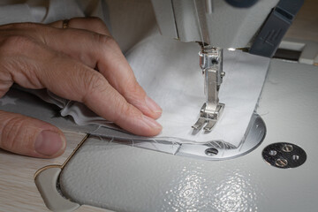 The white fabric is sewn on ar automatic sewing machine.. Woman's hand.