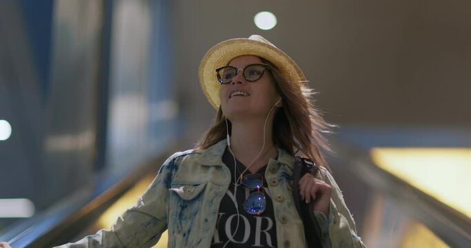 People slow motion 4K. Close up portrait of beautiful smiling woman 30s going down on escalator at night city. Female hipster in stylish outfit, trendy glasses and hat listening music via headphones