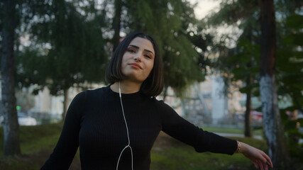 Fototapeta na wymiar Smiling young beautiful woman with in headphones listens to music and dances walking along park slow motion