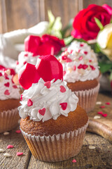 Cupcake with Heart for Valentines. Valentine Day Sweet Dessert, vanilla cupcakes with whipped vanilla cream and red sugar hearts decor for Valentine's Day, wooden background with rose flowers bouquet