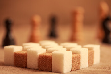 White and brown sugar cubes in form of a chessboard and chessmen in the background .  Closeup. Bokeh. Copy space