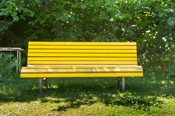 A yellow bench in front of a green wood or shrub. The bench is in the shade under trees. A good cooling off on hot summer days. Time to relax.