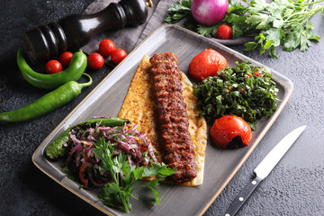 Turkish cuisine. Grilled meat on pita bread with grilled vegetables and herbs. Lula kebab with salad on a grey plate with spices and a knife on a black table. Restaurant menu, horizontal