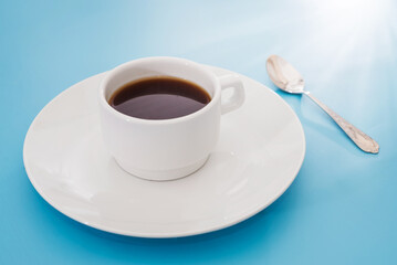 white coffee cup and spoon isolated on blue background. The sun's rays shine on the cup.