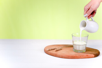 A woman's hand holds a milk jug and pours milk into a clear glass. White and green background. Copy space.