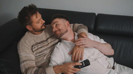 Close up. Two gay men in casual outfits sitting on sofa embracing and watching TV at home. High quality photo