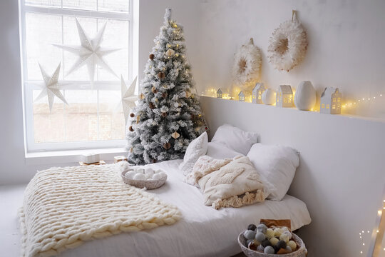 New Years bright interior bedroom with decorated Christmas tree with garlands, white bed, knitted blanket, stairs, stars on window. Christmas interior. Scandinavian style. Location for photo shoot.