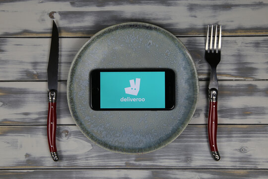 Viersen, Germany - May 9. 2020: Close up of mobile phone screen with logo lettering of food delivery service deliveroo on wood table with dish and cutlery (focus on center)