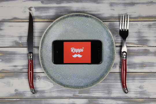 Viersen, Germany - May 9. 2020: Close up of mobile phone screen with logo lettering of food delivery service Rappi on wood table with dish and cutlery (focus on center)