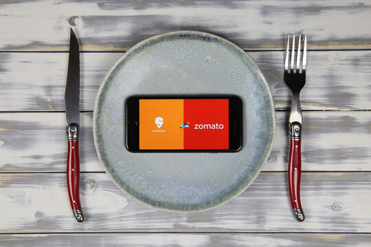 Viersen, Germany - May 9. 2020: Close up of mobile phone screen with logo lettering of indian food delivery service Swiggy Zomato on wood table with dish and cutlery (focus on center)