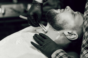 Barber shaving a bearded man in a barber shop. Close up of a hairdresser's hands in black gloves shaving a client's beard