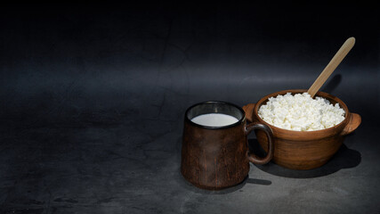 ceramic bowl with cottage cheese, mug with milk on a dark background. Still-life. the style is...
