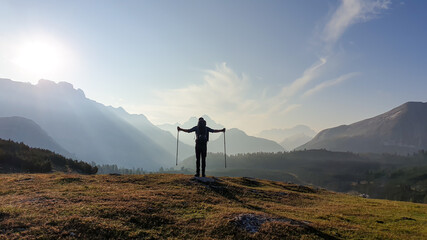 A man with hiking sticks spreading his arms wide open on an early morning in Italian Dolomites. The valley below is shrouded in morning haze. In the back there are high mountain chains. Golden hour