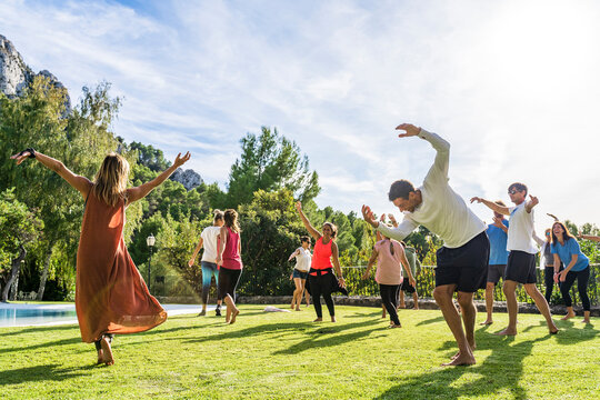 Mature female fitness instructor dancing with tourists at health retreat on grass against sky during sunny day - Powered by Adobe