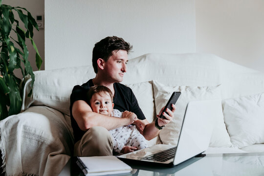 Father text messaging on smart phone holding son while sitting on sofa at home