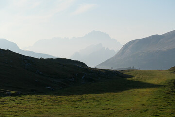 An early morning in Italian Dolomites. Lush green meadow in front. The valley is shrouded in morning haze. In the back there are high mountain chains. Sun slowly rising above the peaks. Golden hour