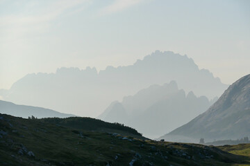 Obraz premium An early morning in Italian Dolomites. Lush green meadow in front. The valley is shrouded in morning haze. In the back there are high mountain chains. Sun slowly rising above the peaks. Golden hour
