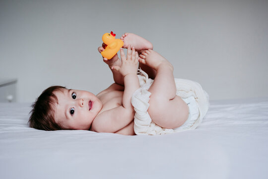 Cute baby boy playing with duck toy while lying down on bed at home