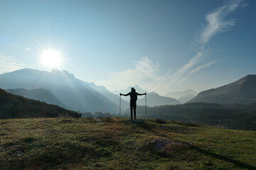 Naklejka premium A man with hiking sticks spreading his arms wide open on an early morning in Italian Dolomites. The valley below is shrouded in morning haze. In the back there are high mountain chains. Golden hour