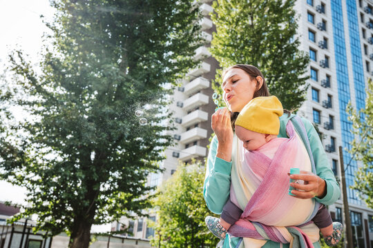 Mother Carrying Baby Blowing Bubble While Standing On Street In City