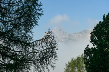 An early morning in Italian Dolomites. A thick forest overgrowing the area. The valley is shrouded in morning haze. There are high, stony mountain chains in the back. Golden hour. New day beginning