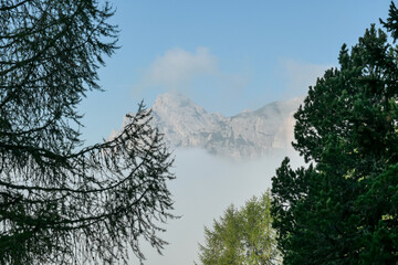 An early morning in Italian Dolomites. A thick forest overgrowing the area. The valley is shrouded in morning haze. There are high, stony mountain chains in the back. Golden hour. New day beginning