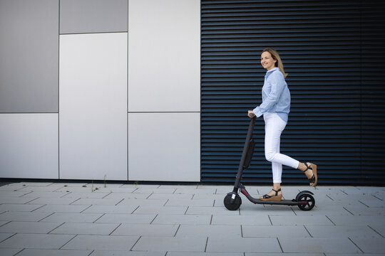 Mid adult woman driving electric push scooter on footpath by metal wall