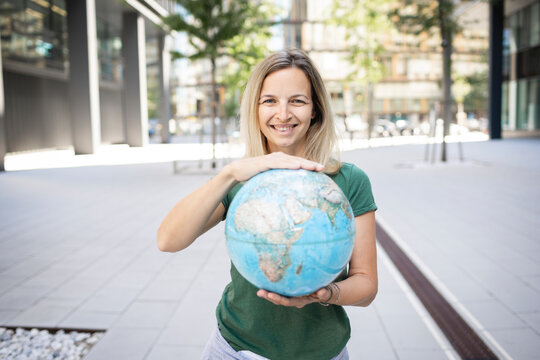 Smiling woman holding globe while standing on footpath in city