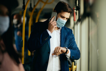 Businessman with face mask talking on the phone while checking time on wristwatch in a bus.