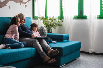 A grandmother with her two granddaughters, reading a book to them on the sofa at home.