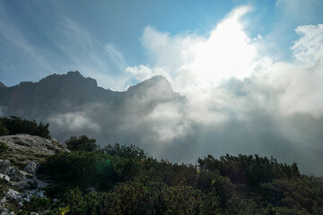 A view on a high and sharp mountains towering above the ground level. The peaks are shrouded with a few clouds. Sunbeams coming through the clouds. Stony, steep mountain walls. Discovering