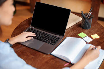 Woman sitting at desk using laptop with blank screen, writing